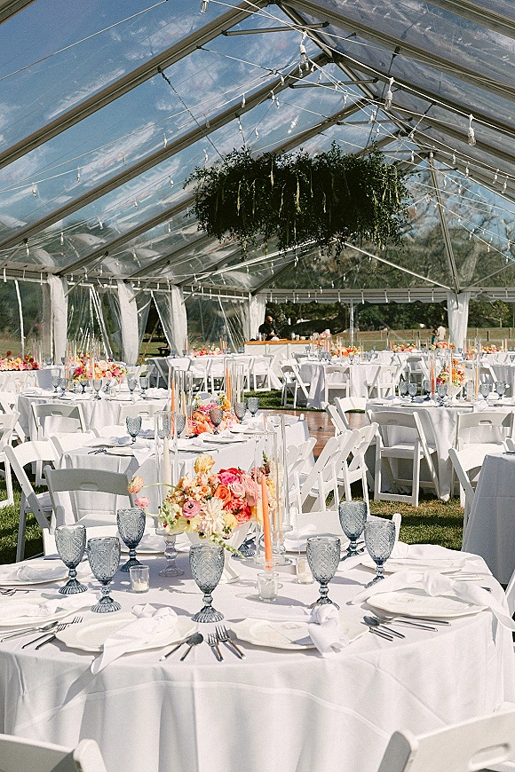 Reception tablescape under a clear top wedding tent with string lights, hanging greenery, floral centerpieces, candles, and blue goblets on white linens