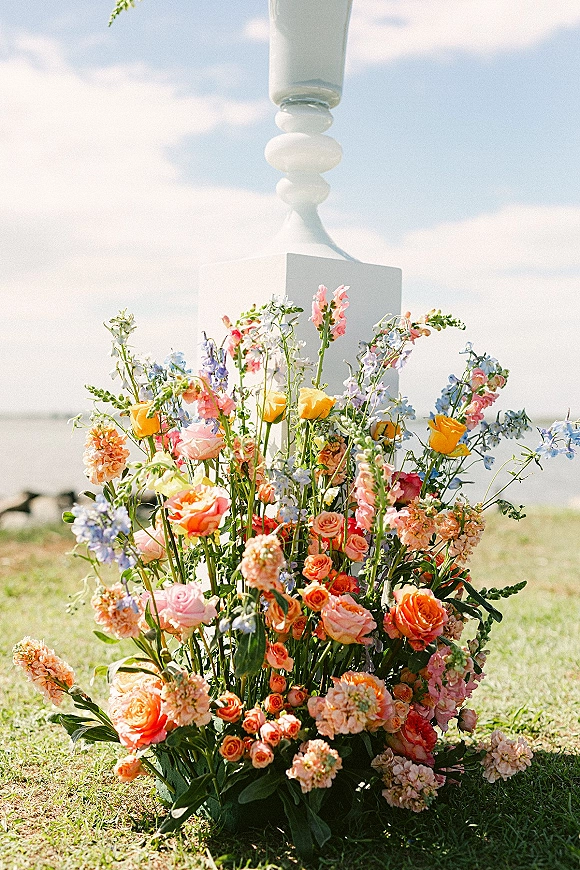 Wedding ceremony florals with a ground floral arrangement of roses and greenery on a white pedestal, set on a lawn with ocean views