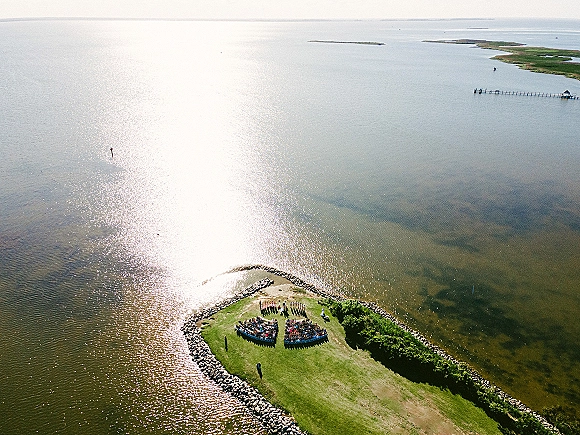 Outdoor wedding ceremony with aerial wedding ceremony view, curved chair rows and aisle runner on a grassy peninsula beside ocean water and rock jetty