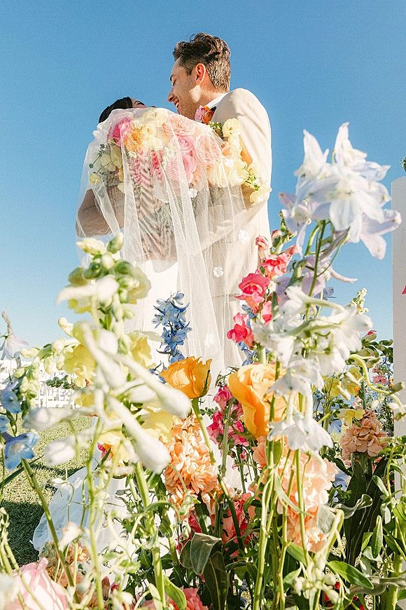 Wedding couple portrait of the groom lifting the bride as they hug, her bridal veil and colorful bouquet framed by blue sky and white chairs