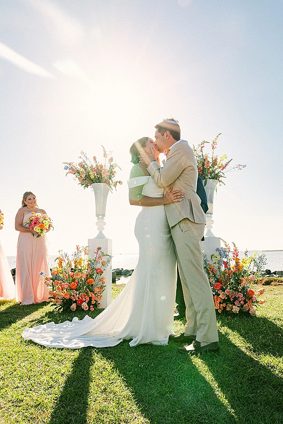 Wedding kiss at a beach wedding ceremony, bride in off-shoulder gown with train and bouquet as sunlit ocean and sky glow behind.