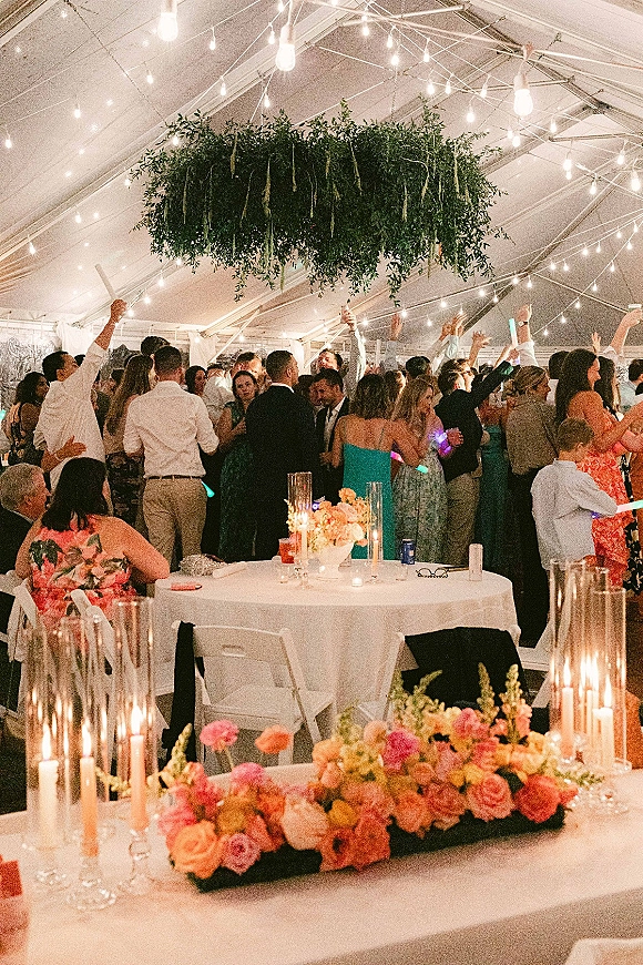 Wedding reception dance floor packed with guests waving glow sticks under string lights and hanging greenery in a clear-top tent