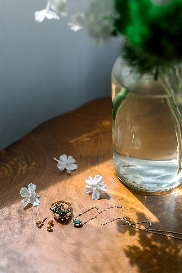 Wedding jewelry flatlay of gold earrings, rings and pendant necklace with white flowers on a wooden table against a blue wall with sunlight shadows