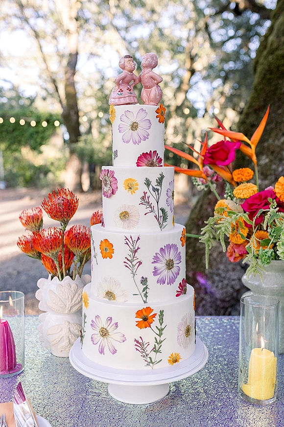 Wedding cake with pressed flower decorations on a pedestal stand, topped with figurines, beside candles and bright florals under string lights