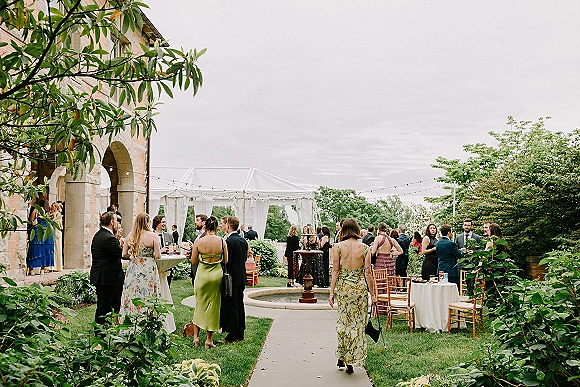Cocktail hour with wedding guests mingling around high-top tables and welcome drinks under string lights in a clear tented garden courtyard