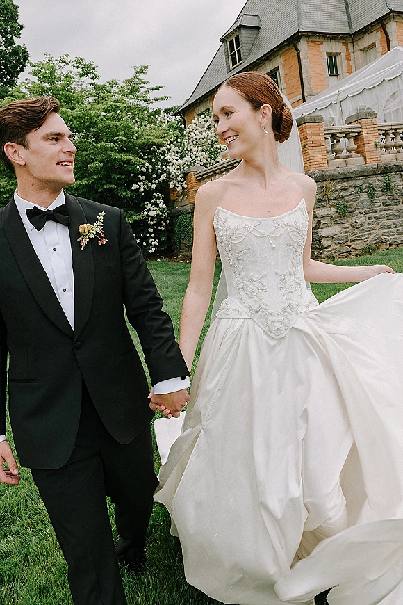 Couple portrait of bride and groom holding hands, bride in strapless gown and veil with earrings, beside stone wall and garden lawn
