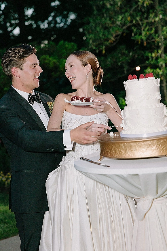 Wedding cake cutting as bride and groom share a bite, laughing beside a raspberry-topped tiered cake under bistro lights in a garden night setting