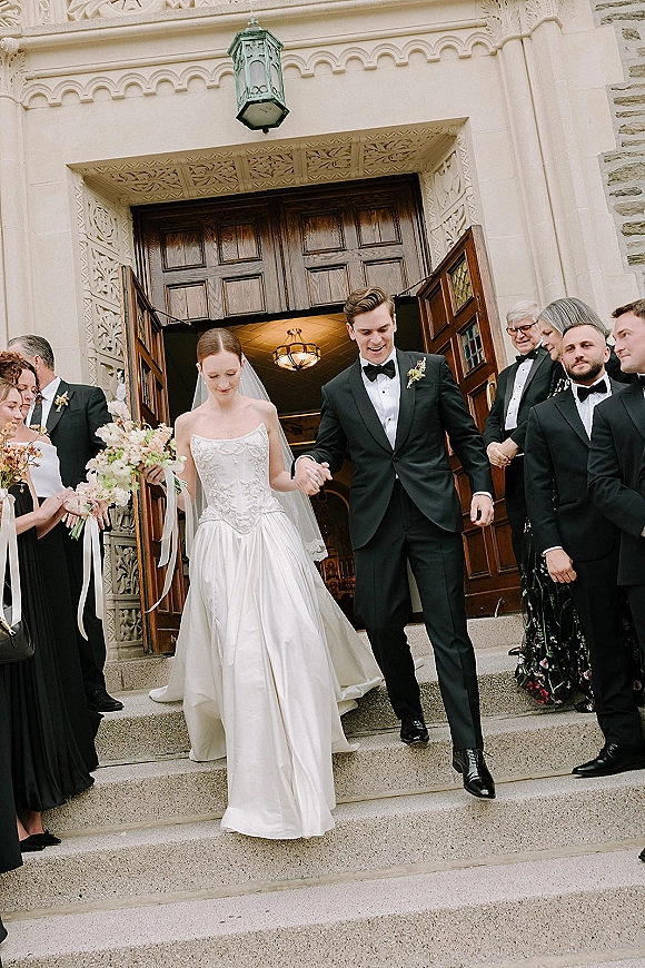 Wedding recessional as bride and groom exit a stone church, holding hands on steps, bouquet ribbon streamers, veil, and tuxedoed party behind
