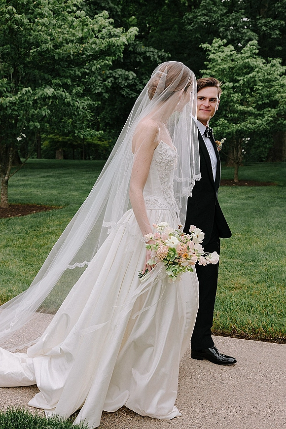Couple portrait of bride and groom walking on a garden walkway, bride in strapless lace dress with cathedral veil, holding pastel bouquet