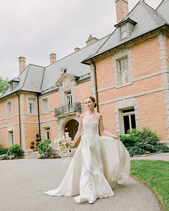 Bridal portrait of a bride in a strapless wedding dress holding a pastel bouquet with trailing ribbons on a mansion driveway under overcast sky