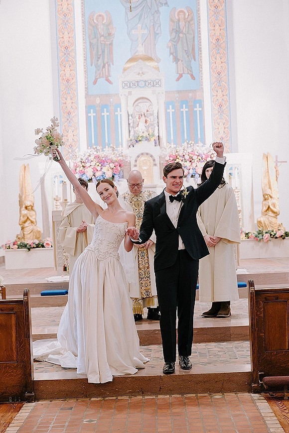 Recessional moment as bride and groom celebrate, bouquet in hand and fist raised, walking down the church aisle past the altar flowers