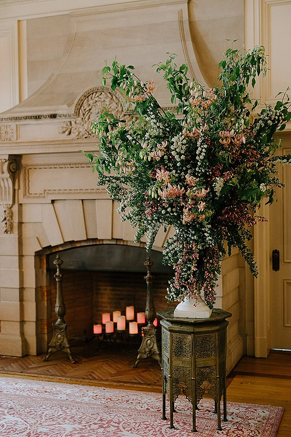 Wedding floral arrangement in a large wedding centerpiece with branchy greenery and blush blooms in a white vase before an ornate fireplace mantel