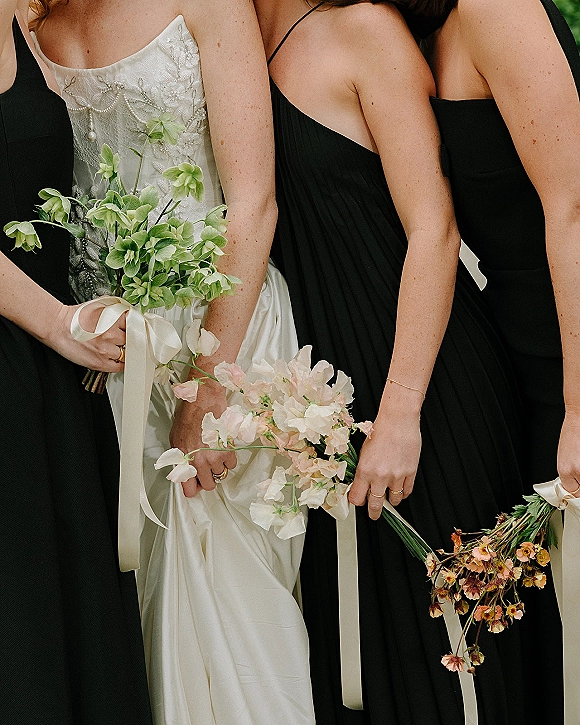 Bridesmaid bouquets in black bridesmaid dresses, mixed blooms with long ribbon wraps held beside a lace bridal gown amid greenery outdoors