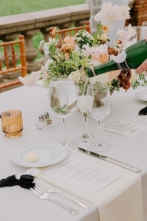 Reception tablescape wedding table setting with white tablecloth, floral centerpiece, champagne bottle and glassware on a stone terrace overlooking lawn