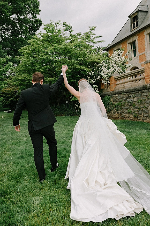 Wedding couple portrait of bride and groom walking away hand in hand, her veil and long train flowing on a green lawn by a brick manor