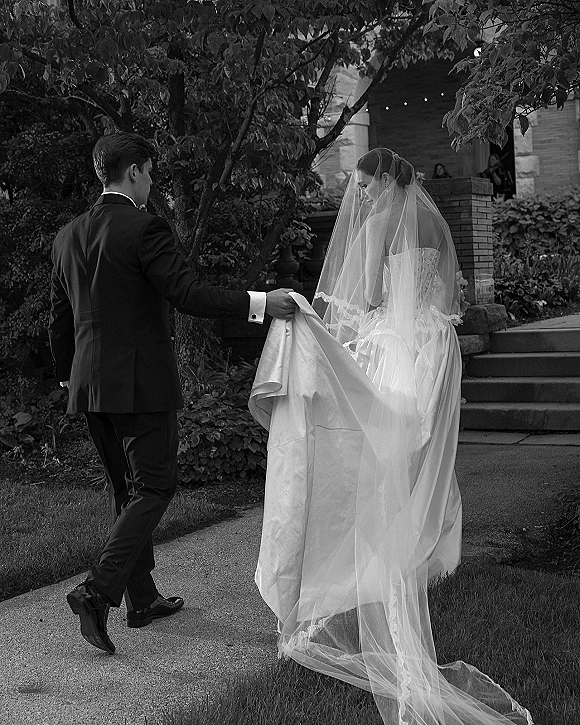 Wedding couple portrait with groom in tuxedo lifting the bride’s long veil and dress train on stone steps by a brick entryway with string lights