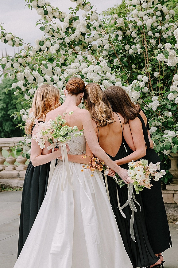 Bridesmaid group photo with bride and bridesmaids hugging, holding bouquets with ribbon streamers on a garden terrace by a stone balustrade