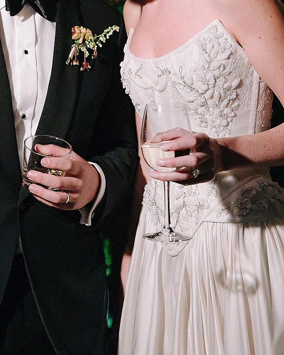 Wedding reception toast as bride and groom raise wine and whiskey glasses, beaded bodice and tuxedo details glowing in dark lighting