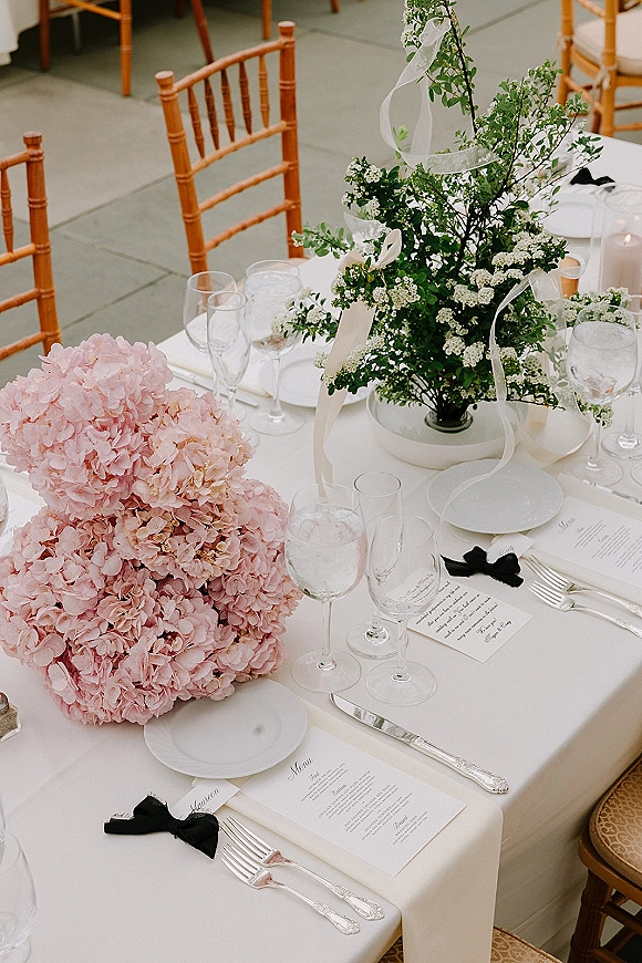 Reception tablescape wedding table setting with pink hydrangea and greenery centerpieces, white plates, menus, and gold chiavari chairs outdoors