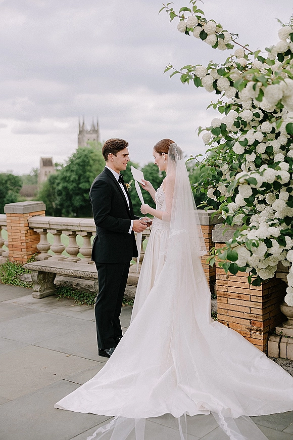 Wedding vows as bride reading vows to groom, holding vow cards on a stone terrace by balustrade under cloudy sky with veil train