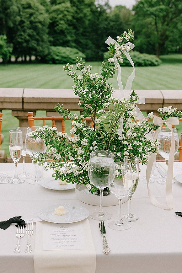 Reception tablescape with a wedding table centerpiece of white flowers and greenery branches, set with silver flatware on a stone terrace lawn backdrop