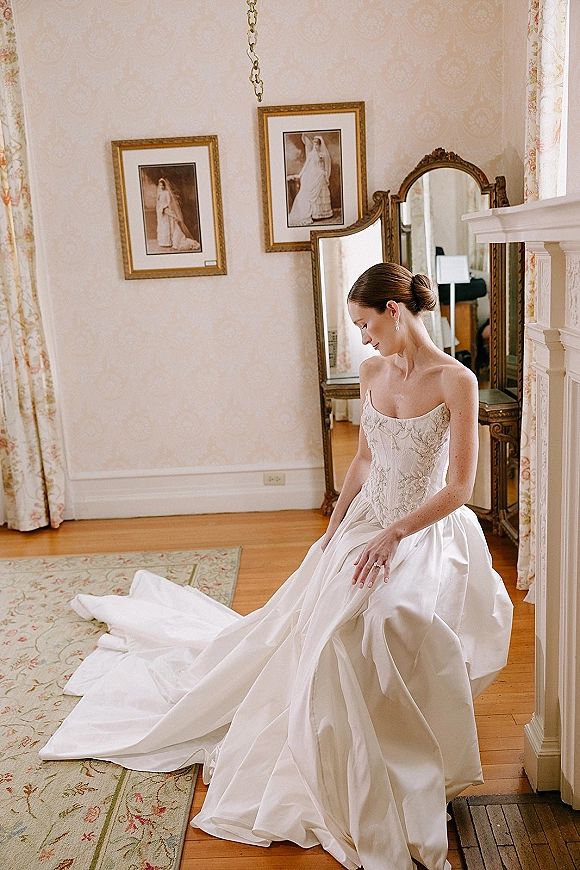 Bridal portrait of a getting ready bride in a strapless embroidered gown with long train and pearl earrings in a vintage room by a fireplace mantel