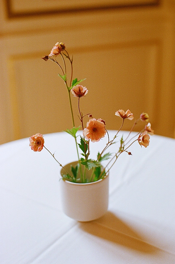 Bud vase centerpiece with peach flowers in a white ceramic vase on a round table, lit by sunlight against wooden wall paneling