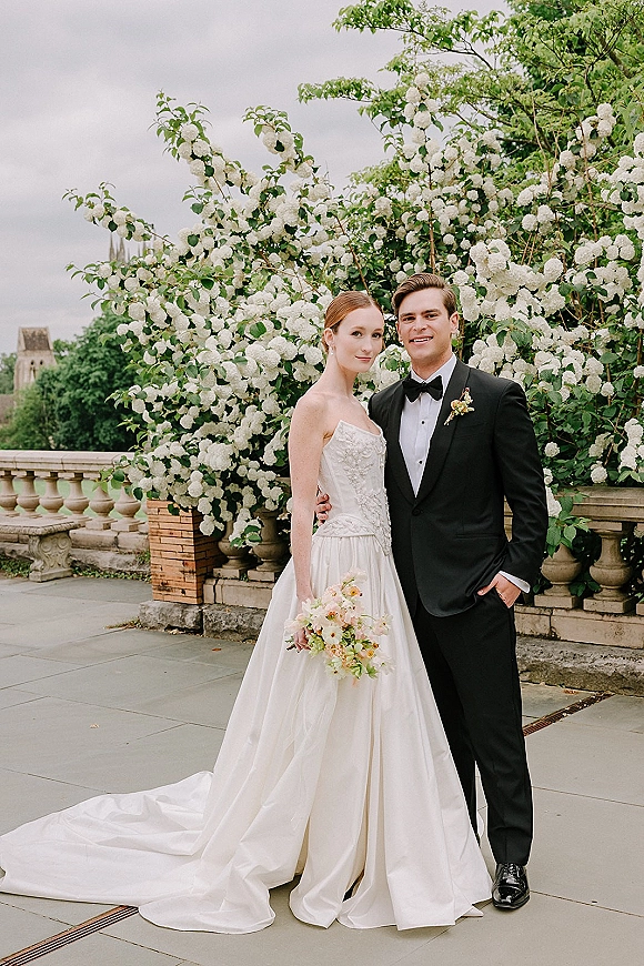 Couple portrait of bride and groom portrait on a terrace by a stone balustrade, bride holding a pastel bouquet before flowering bushes