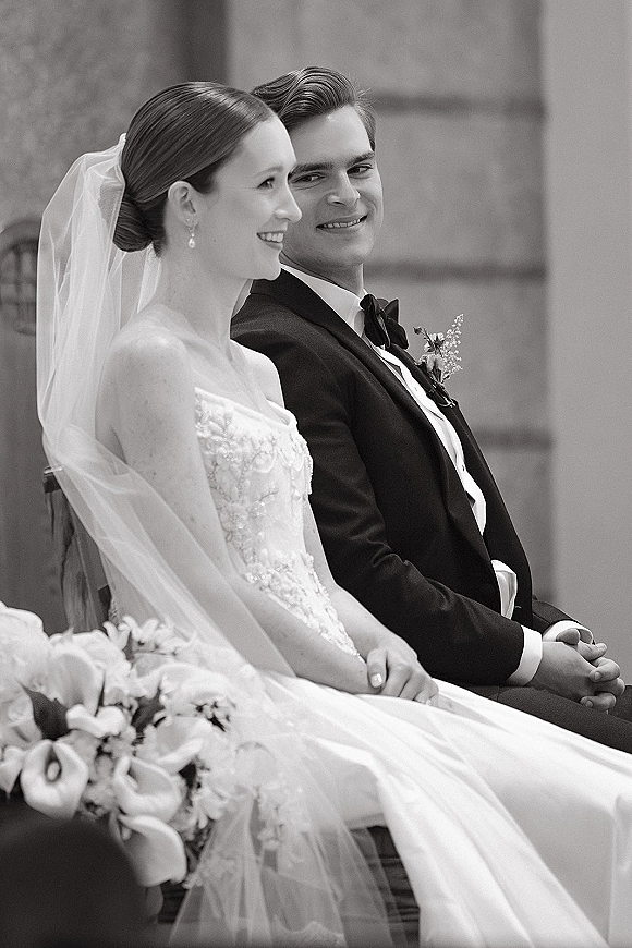 Bride and groom in a black and white wedding portrait seated by a stone wall, groom gazing at her as she smiles holding bouquet