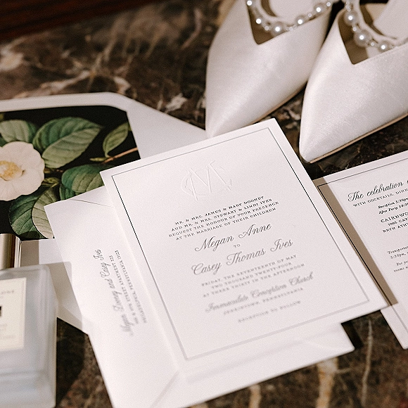 Invitation suite with letterpress wedding invitations, RSVP card and envelopes beside pearl bridal shoes, greenery and wax seal on marble tabletop