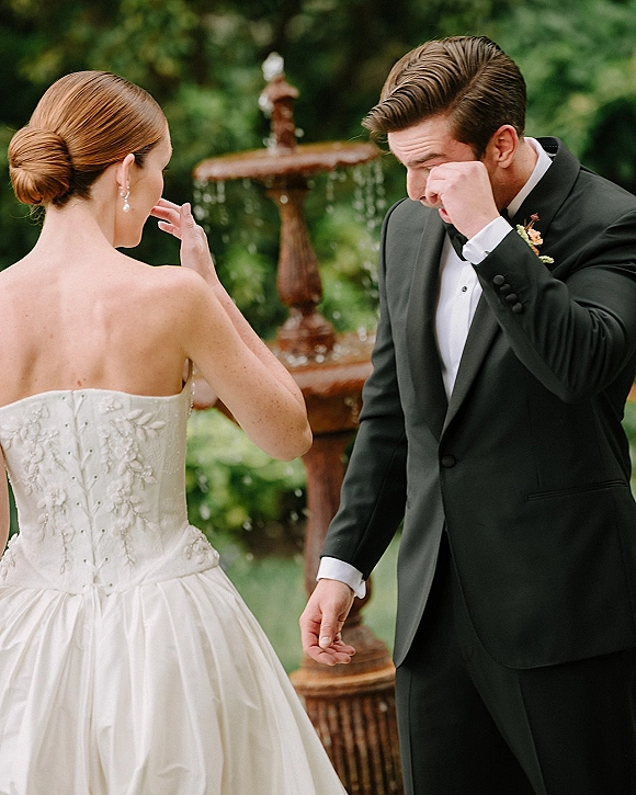 First look moment as bride in a strapless wedding dress comforts groom wiping tears in a tuxedo beside a garden fountain