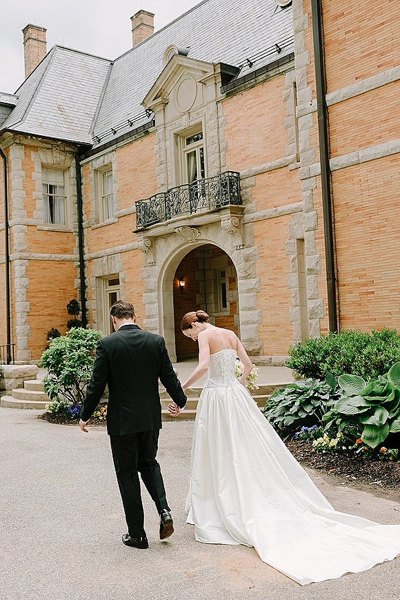 Couple portrait of newlyweds holding hands as they walk away, bride’s long train and bouquet flowing on steps by a brick archway