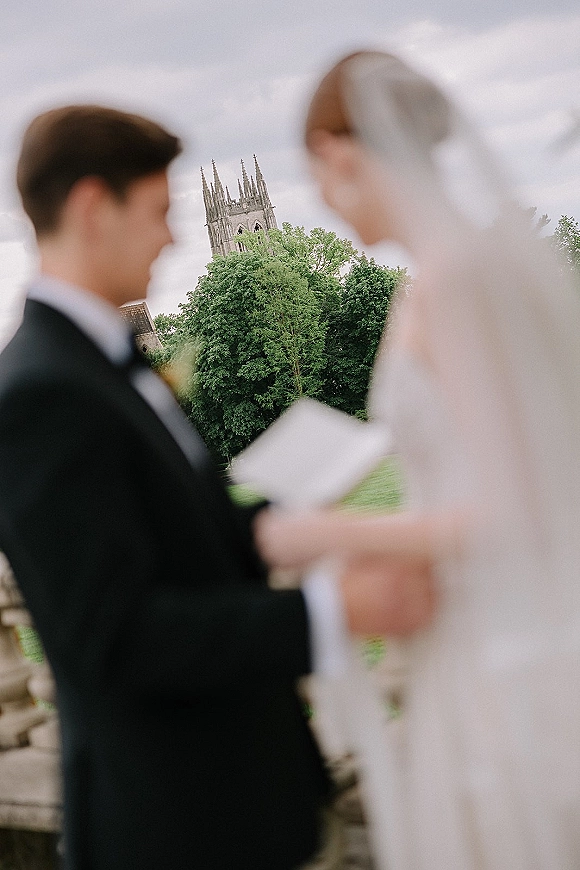 Wedding vows as bride and groom reading vows from vow books on a stone terrace, her long veil in the breeze with church spires behind