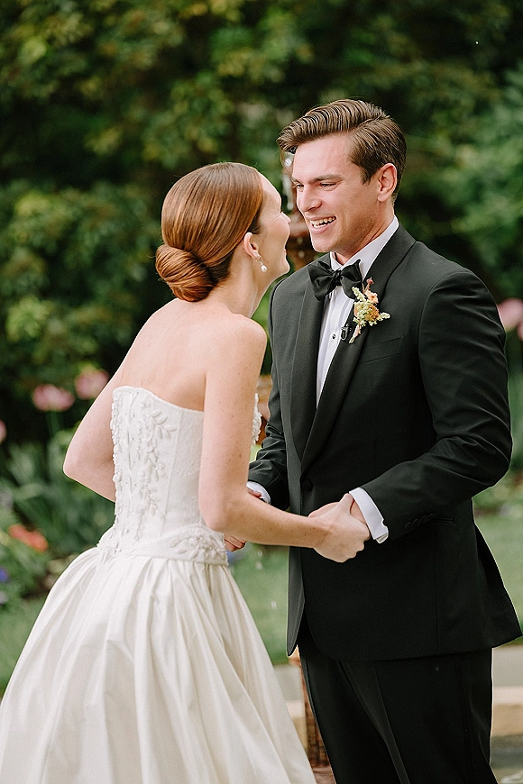 First look moment as bride and groom hold hands, her strapless dress and pearl drop earrings against soft garden greenery backdrop
