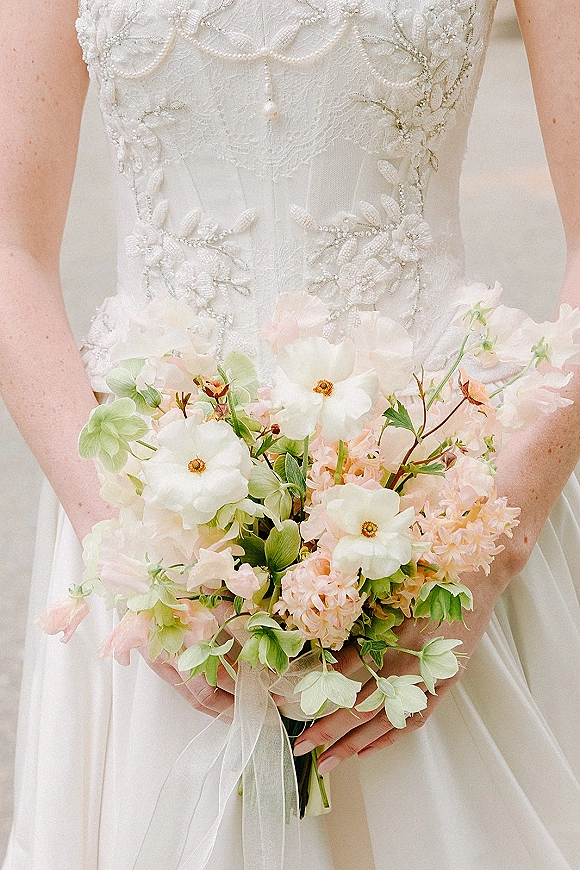 Bridal bouquet of white and blush flowers with greenery and sheer ribbon held against a strapless beaded lace wedding dress bodice