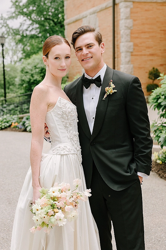 Couple portrait of bride in a strapless floral applique gown holding a pastel bouquet beside groom in black tuxedo on a garden walkway by brick building