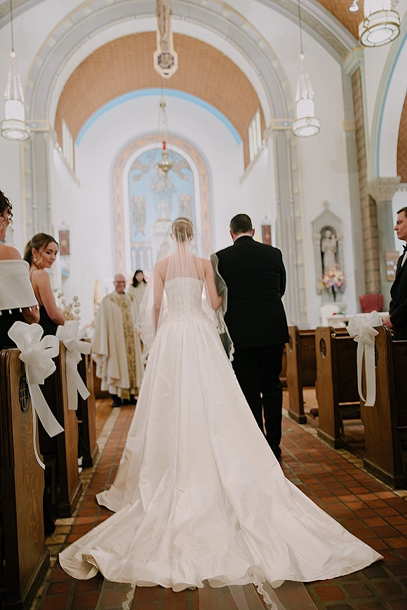 Wedding processional with bride walking down aisle in a strapless gown and long veil, train trailing past pew bows toward the altar