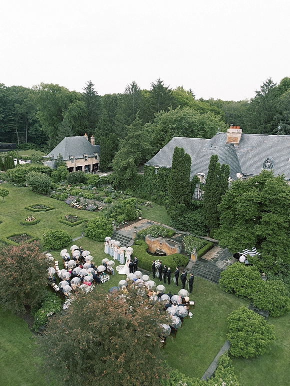 Outdoor wedding ceremony in a garden wedding ceremony, guests hold clear umbrellas as the wedding party lines the floral aisle on a stone terrace