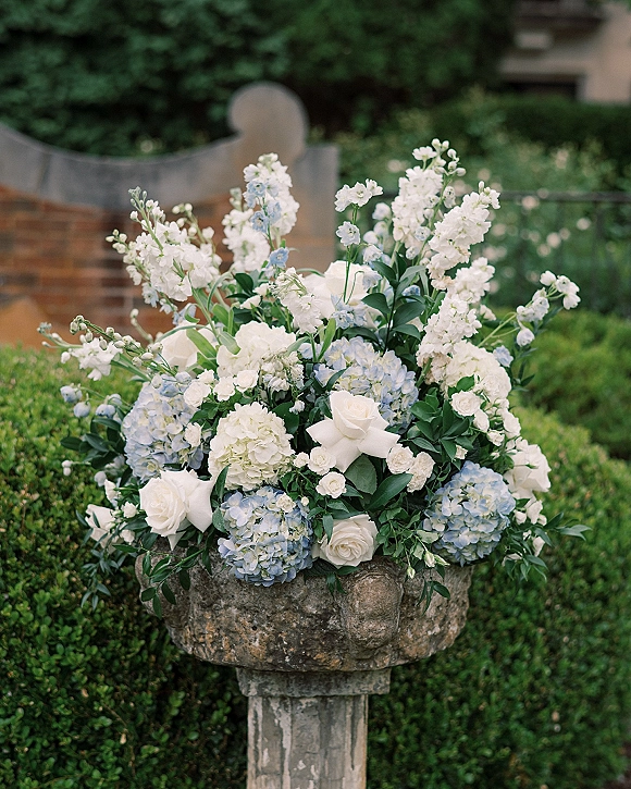 Wedding floral arrangement of white roses and blue hydrangeas with greenery in a stone pedestal urn beside garden hedges and steps