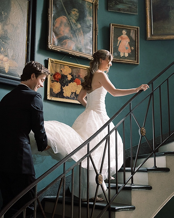Wedding couple portrait of bride walking upstairs as groom holds her dress train on an indoor staircase with teal wall and gold-framed paintings
