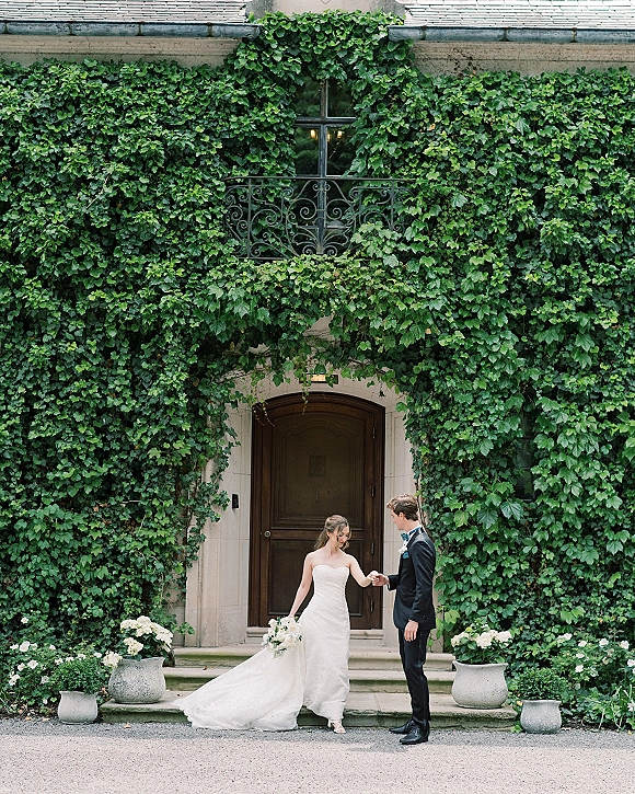 Couple portrait of bride and groom holding hands, bride in strapless gown with long train and bouquet by an ivy-covered arched doorway