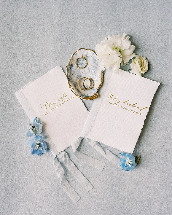 Wedding vow books with gold foil lettering and ribbon ties beside gold rings on a marbled dish with flowers on gray fabric flat lay