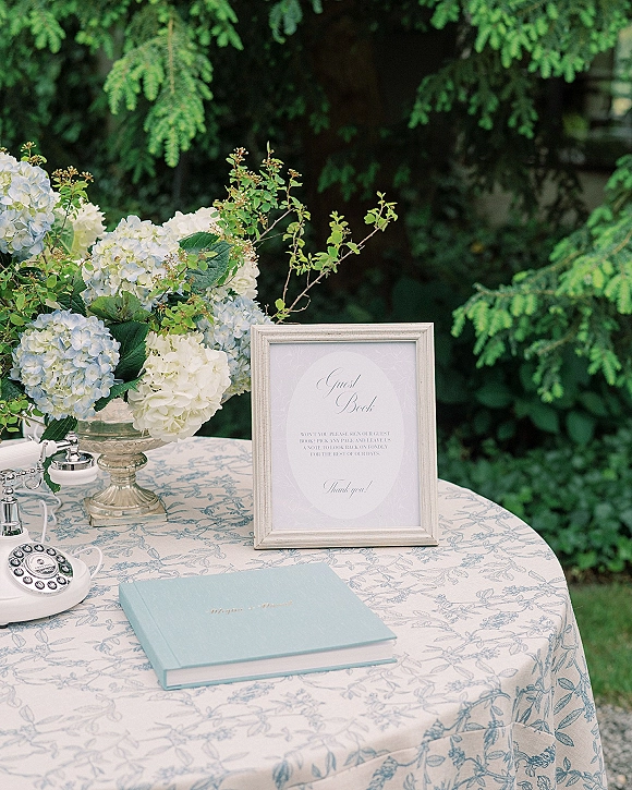 Wedding guest book table with a guest book sign, blue-and-white hydrangea in a glass pedestal vase, and vintage rotary phone amid garden greenery