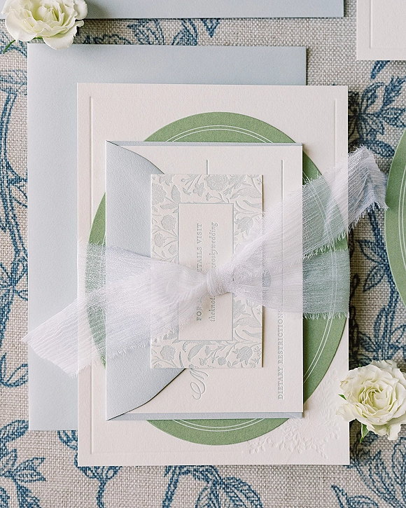Invitation suite with letterpress wedding invitations, RSVP card, vellum ribbon, and envelopes styled with white roses on blue patterned fabric