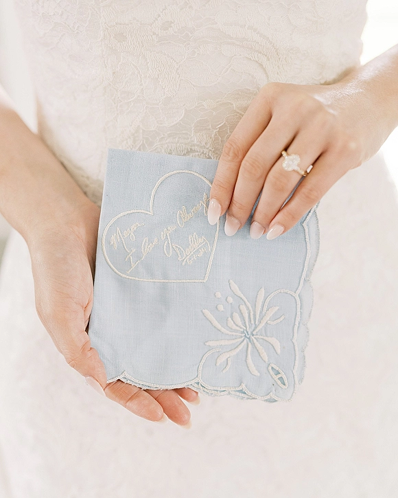 Wedding handkerchief held in bride’s hands, embroidered wedding handkerchief with lace dress detail, engagement ring, and neutral manicure on white backdrop