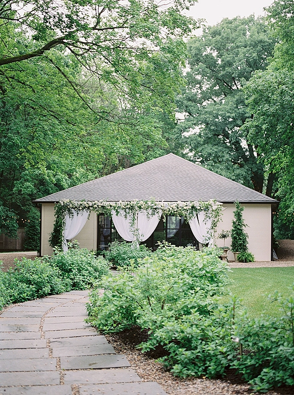 Outdoor ceremony backdrop with draped wedding backdrop in white fabric, floral garland and greenery accents, string lights in a garden pavilion setting