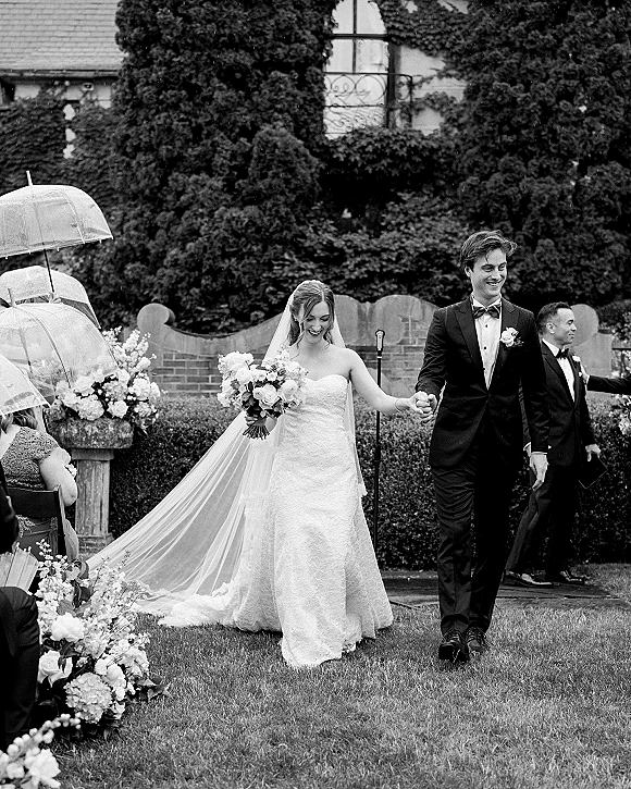 Wedding recessional with bride and groom recessional holding hands under clear umbrellas, bouquet and veil flowing past garden hedge and brick wall