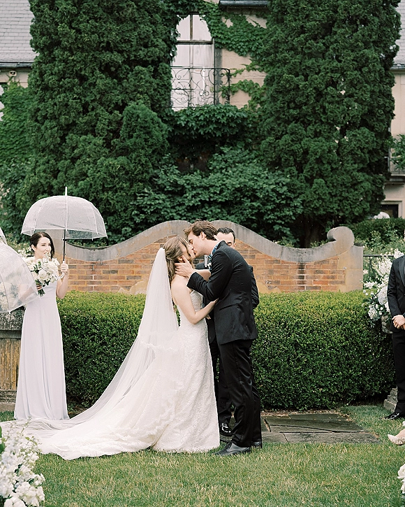 Wedding kiss under a clear umbrella as the bride’s long veil flows and the groom in a tuxedo leans in, with hedges and an ivy brick wall behind