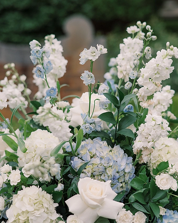 Wedding floral arrangement with blue and white wedding flowers, white roses and hydrangea, greenery accents, set against garden greenery and a blurred stone feature