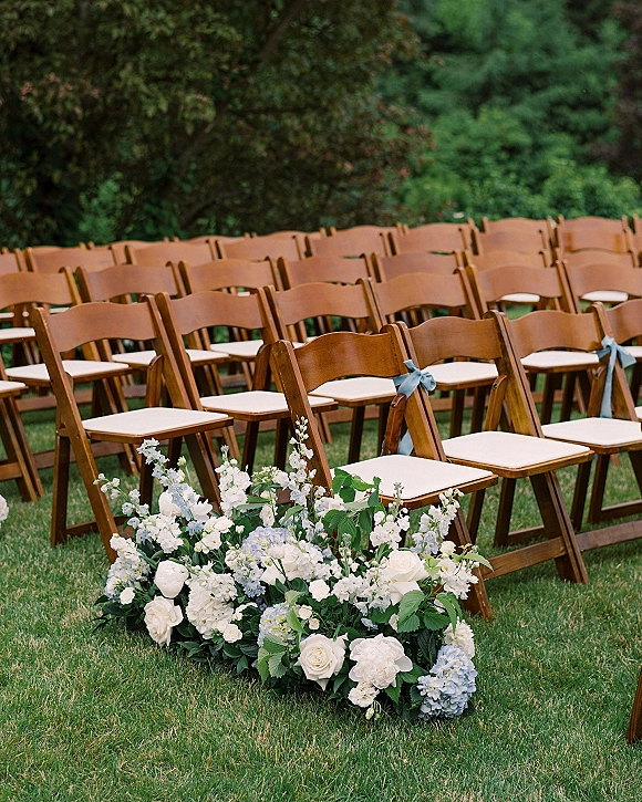 Ceremony seating with outdoor wedding chairs, white cushions and blue ribbon bows lining a grassy garden aisle with hydrangeas and roses
